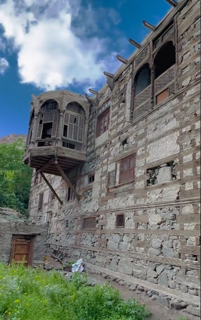 Ancient Kharmang Fort featuring stone walls and a traditional carved wooden balcony overlooking a grassy courtyard under a blue sky in Gilgit-Baltistan, Pakistan.