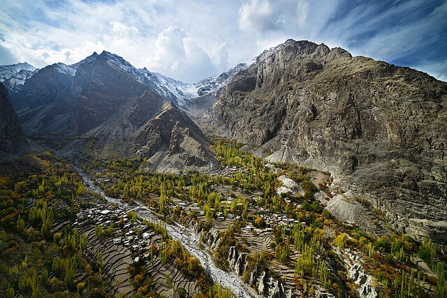 High-angle scenic view of golden autumn trees and terraced village fields in Khaplu Valley, Ghanche, Gilgit-Baltistan, Pakistan, framed by massive rocky Karakoram mountains with snow-capped peaks.