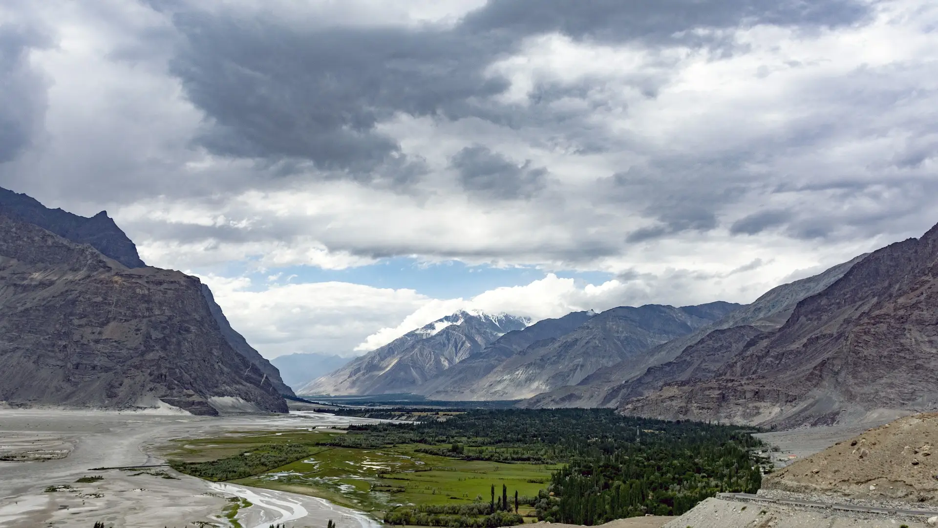 Lush green Shigar Valley oasis with a winding river surrounded by rugged Karakoram mountains under a dramatic cloudy sky in Gilgit-Baltistan Pakistan.