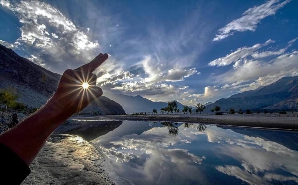 A dramatic sunrise reflecting over a calm pool of water in Shigar Valley, Skardu, Gilgit-Baltistan, Pakistan, with a traveler’s hand framing the distant mountain peaks.