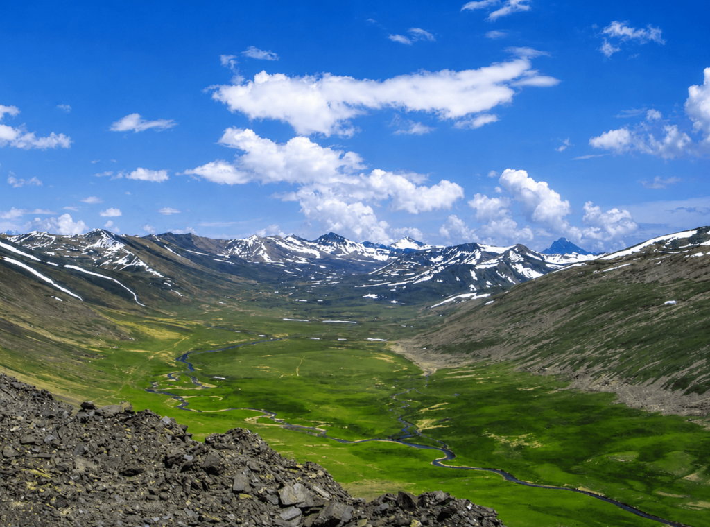 Scenic view of Babusar Top mountain valley with green meadows, winding stream, and snow-capped peaks