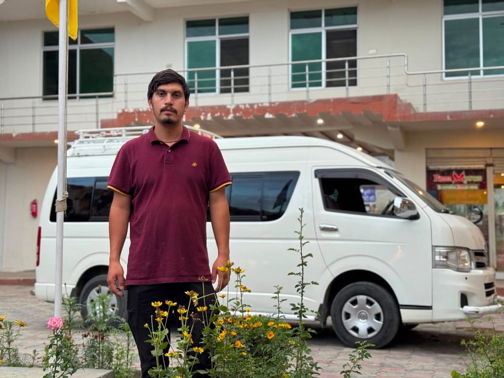 Young man standing in front of a white passenger van outdoors, with flowering plants in the foreground and a building in the background.