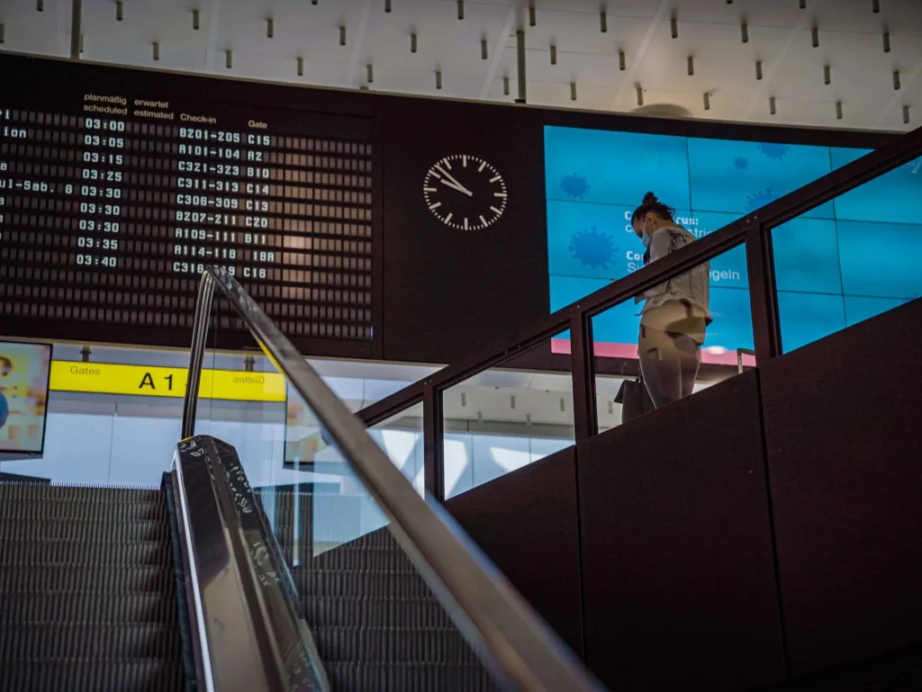 Traveler on an escalator inside an airport departure terminal with a flight information board and departure gates in the background.