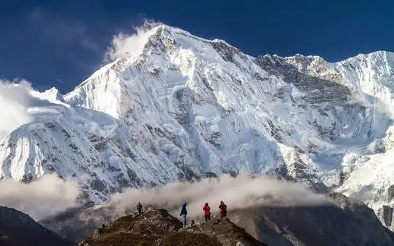 Trekkers standing on a mountain viewpoint overlooking a towering snow-covered Himalayan peak beneath a clear blue sky.