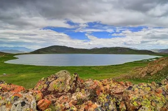 High-altitude lake surrounded by green plains and rocky mountains under a cloudy sky