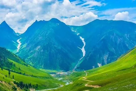Lush green meadows and winding tracks leading through Minimarg in Astore Valley, Gilgit-Baltistan, framed by majestic snow-dusted mountains and dramatic clouds.