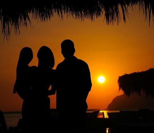 Silhouette of a family of three, with a mother holding a child standing beside a father on a beach at sunset under a thatched umbrella roof, looking out at the golden sun setting over the ocean.