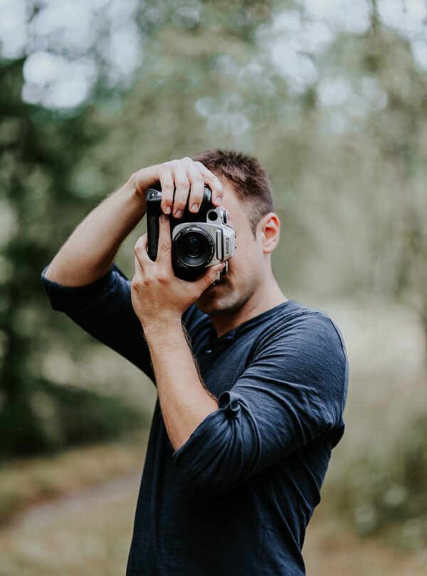 A male photographer wearing a dark blue long-sleeve shirt holding up a vintage-style Canon SLR camera to his eye, blocking most of his face, set against a soft bokeh forest background.